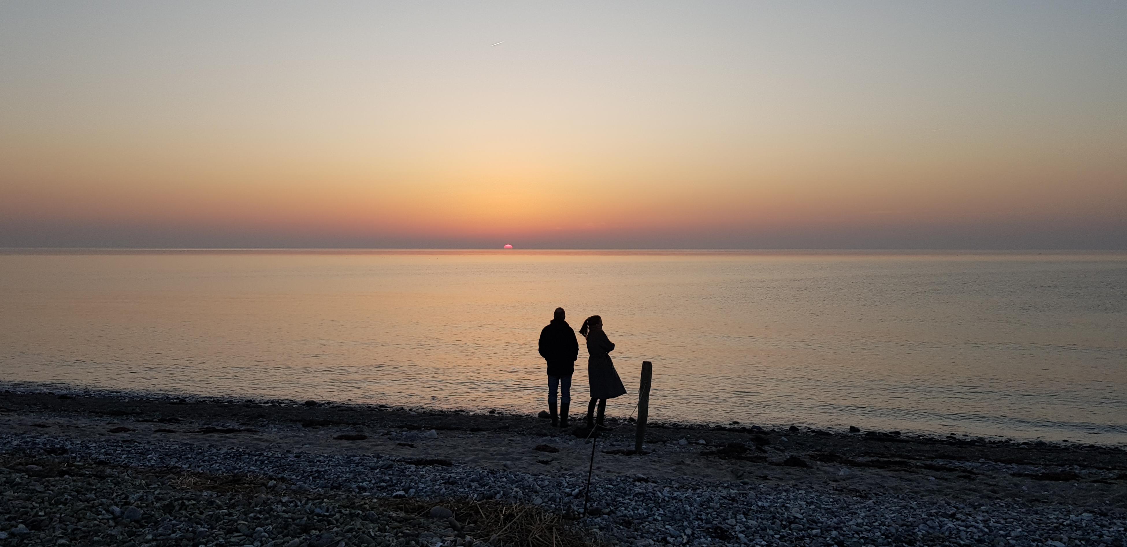 Zwei Personen stehen als schwarze Silhouetten an einem steinigen Ufer.
Die linke Person hat eine eher männliche Statur, steht mit dem Rücken zum Fotografen, hat die Hände in den Hosentaschen und schaut hinaus aufs Wasser zur untergehenden Sonne, die ein blassrosa Licht über die ansonsten eher graue Szene schickt.
Rechts daneben steht mit etwas Abstand eine Person mit weiblicher Statur. Durch eine Drehbewegung nach rechts schwingen Pferdeschwanz und Mantel. Die Arme sind vor dem Körper verschränkt.
Der Himmel ist klar mit einem nebligen Streifen über dem Horizont. Das Wasser ist fast unbewegt. Das Ufer liegt schon im Dunkeln.