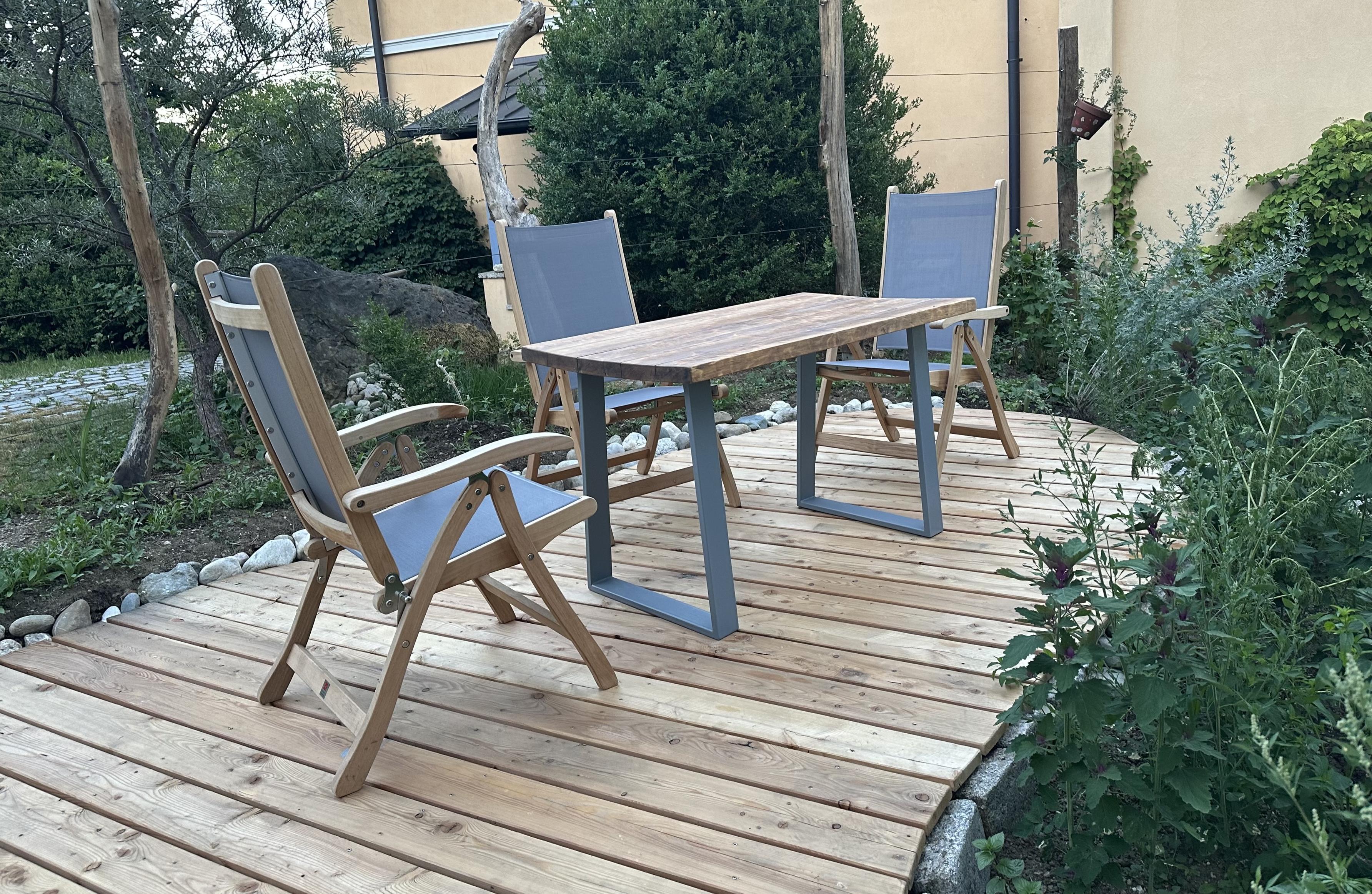 Blick auf eine organisch geformte Terrasse aus frischem Holz. Darauf drei Gartenstühle und ein Tisch, dessen Kufen das gleiche Blaugrau haben wie die Sitzflächen und Lehnen der Stühle